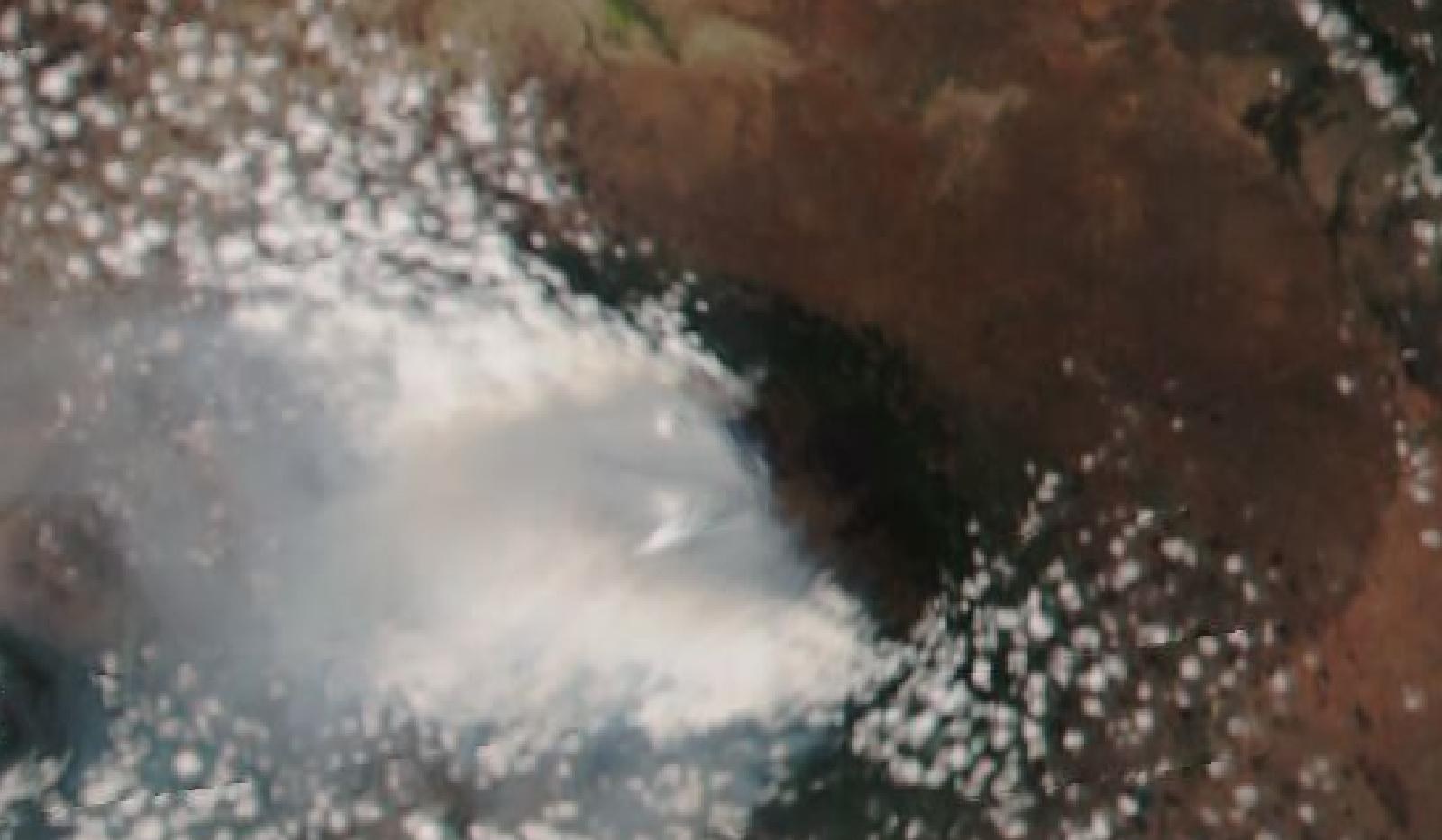 衛星から見た真色画像。画面中央やや右の暗い緑色の山体から、灰白色の煙の帯が左（西）へ広く流れている。周囲は褐色の乾いた大地と白い雲