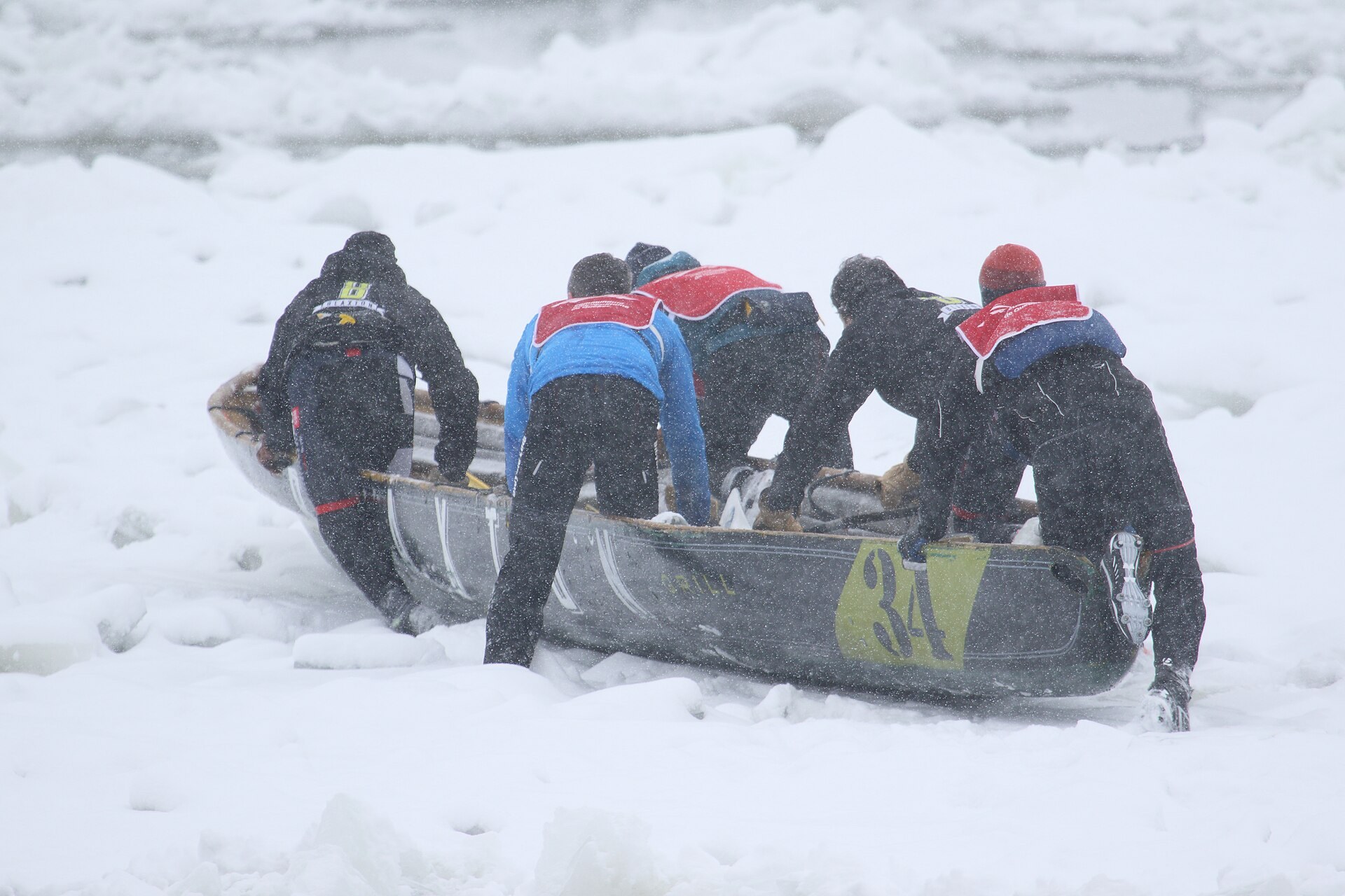雪の降る流氷の上で、五人のチームが番号34のカヌーを押しながら走っている。艇の周りは砕けた氷と雪に覆われている