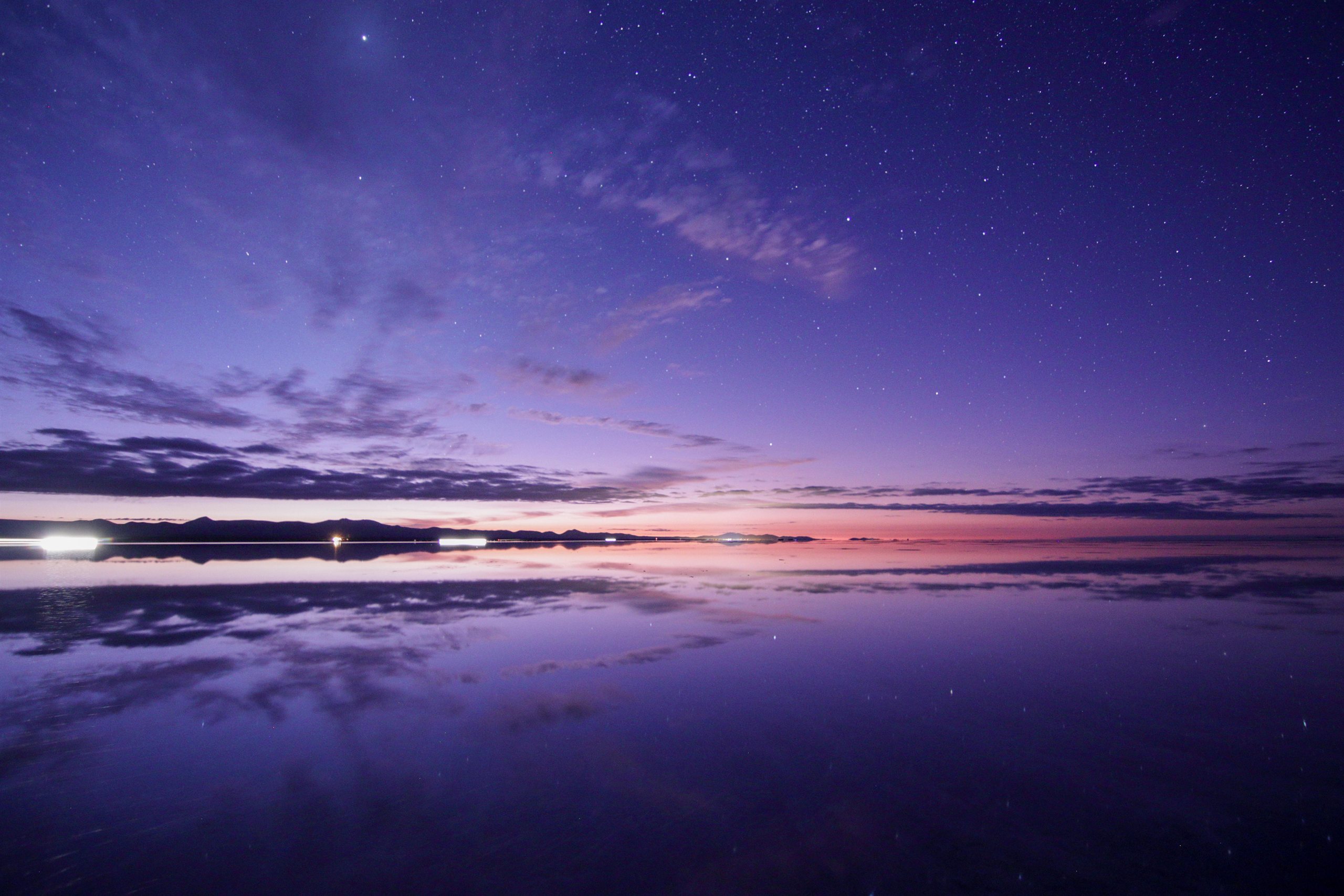 薄紫の薄明が広がる塩湖の水面に、空の雲と星がそのまま映り込み、上下対称に伸びる眺め
